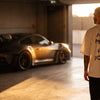 Homme de dos en t-shirt Need Money For Porsche dans un garage, une 911 GT3 RS à contre-jour derrière lui