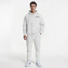 Sweat à capuche blanc pour homme, modèle "Édition Neige" de la marque Tourismo Clothing. Hoodie sans cordon, coupe droite, capuche épaisse et poche kangourou. Logo brodé noir "TOURISMO" avec motif damier placé côté cœur. Le mannequin est debout en studio, de face, main gauche dans la poche, main droite relâchée. Ensemble blanc intégral avec pantalon ajusté et sneakers Nike blanches. Fond blanc minimaliste. Vêtement streetwear premium inspiré du racing, alliant pureté du design et esthétique technique.